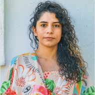 Angelica Monteiro wears a bright patterned top with long curly brown hair pulled to one side.