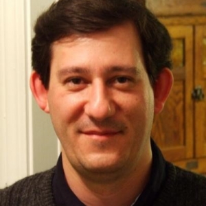 A man with black hair stares at the camera. He is wearing a black collared shirt and dark grey sweater.